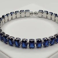 Deep blue emerald-cut sapphire tennis bracelet worn on a woman's wrist against a minimalist, neutral studio background.