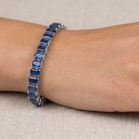 Deep blue emerald-cut sapphire tennis bracelet worn on a woman's wrist against a minimalist, neutral studio background.